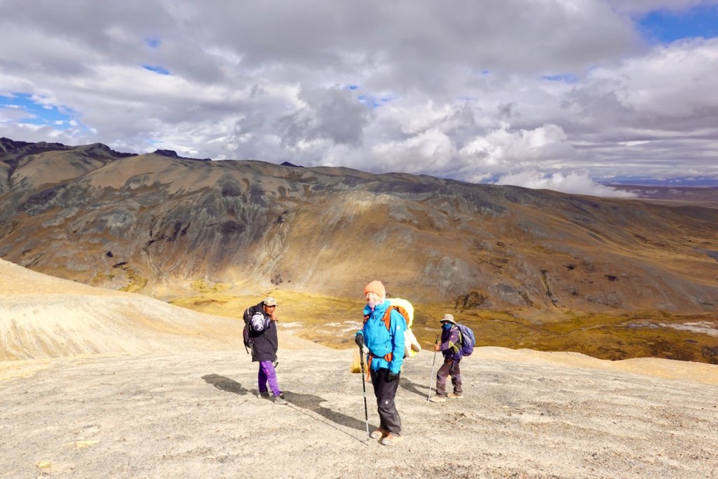 Three hikers navigating a rocky terrain on a mountain with a wide valley and clouds in the background.