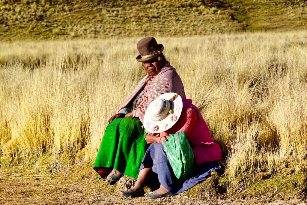 Two women wearing traditional clothing sitting on the ground in a grassy field.