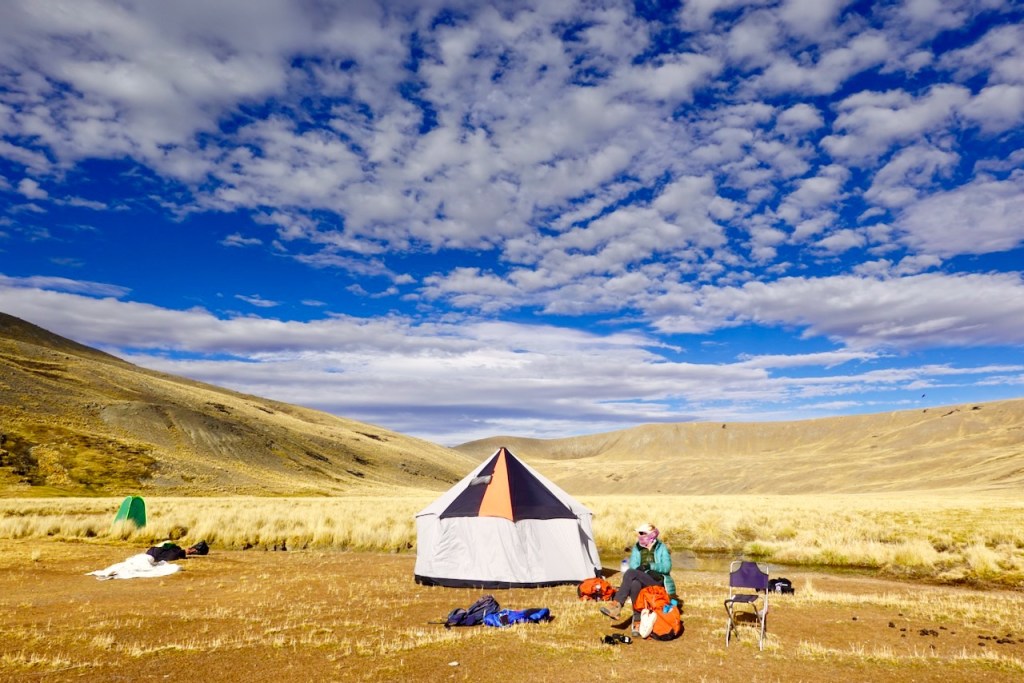 Camping scene in a grassy field with a large tent, surrounded by mountainous terrain and a blue sky filled with clouds. A person is seated nearby with camping gear spread out.