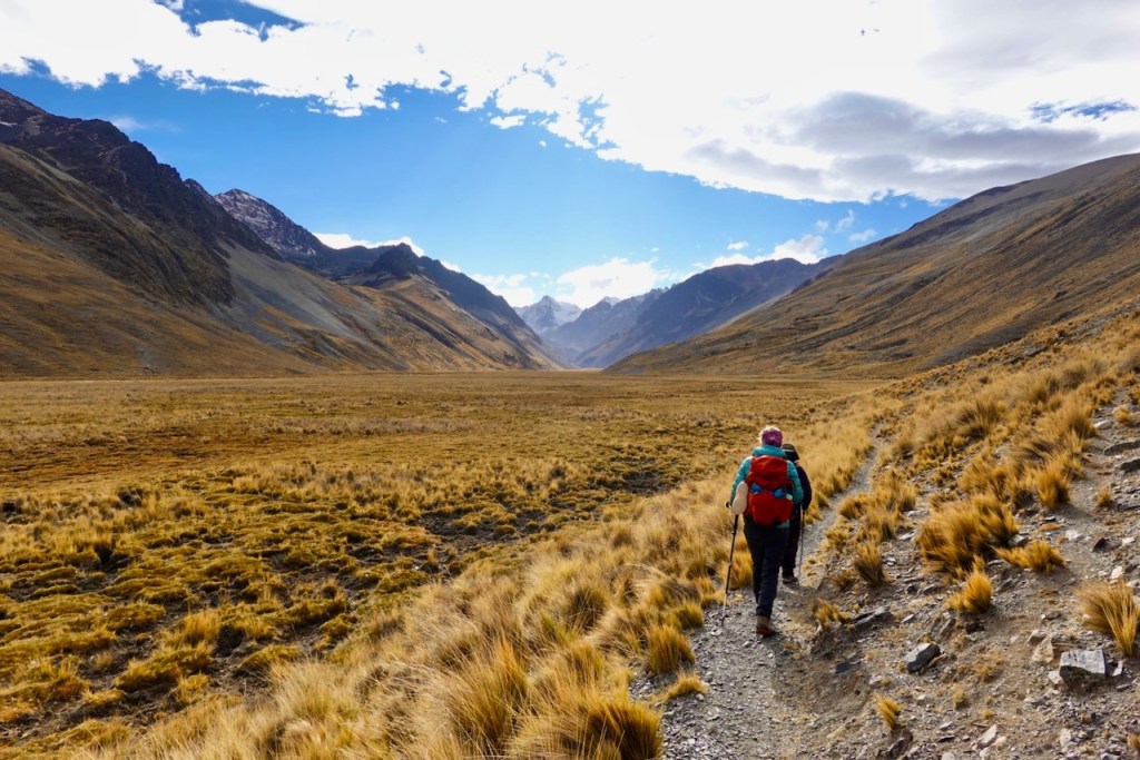 A hiker walking on a narrow trail through a vast grassy valley surrounded by mountains under a blue sky with scattered clouds.
