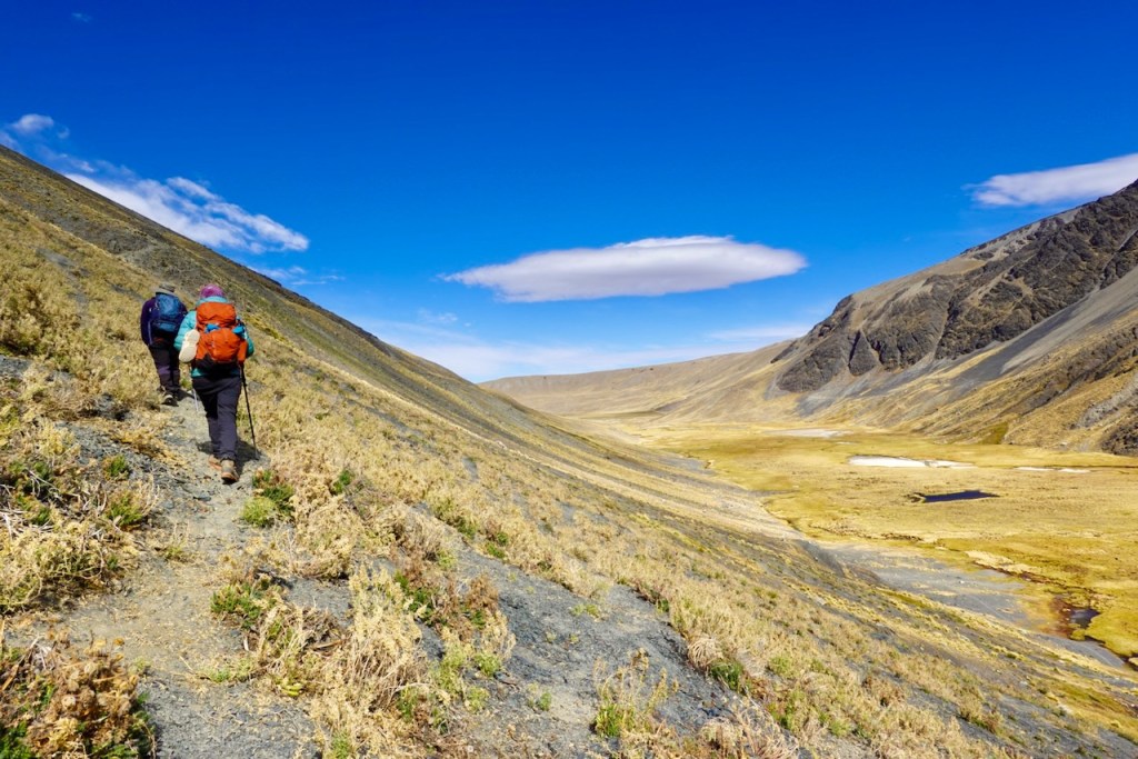 Two hikers walking along a trail in a mountainous landscape under a blue sky with scattered clouds.