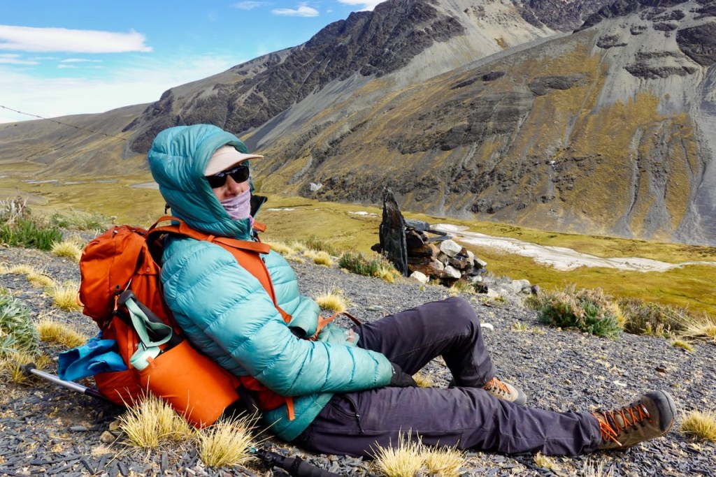 A person wearing a teal puffy jacket and sunglasses, sitting on rocky terrain with an orange backpack, surrounded by a mountainous landscape and grassy patches.
