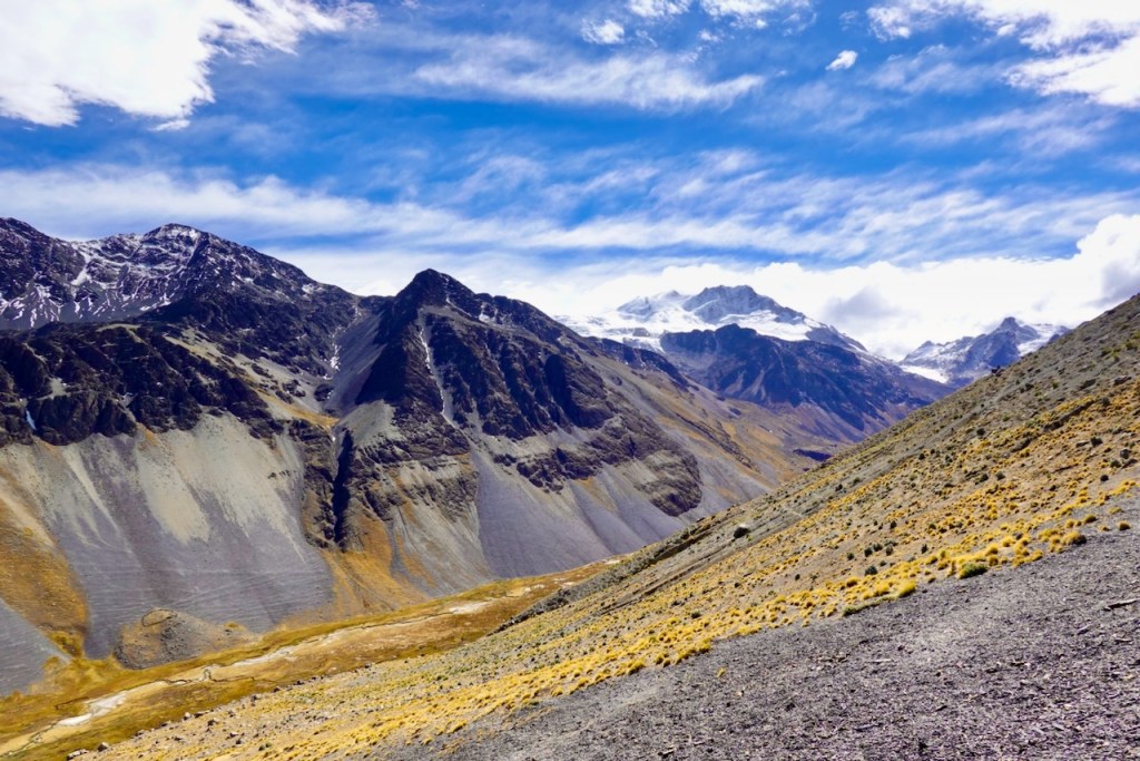 A panoramic view of rugged mountains under a partly cloudy sky, showcasing snow-capped peaks and rocky slopes with sparse vegetation in the foreground.