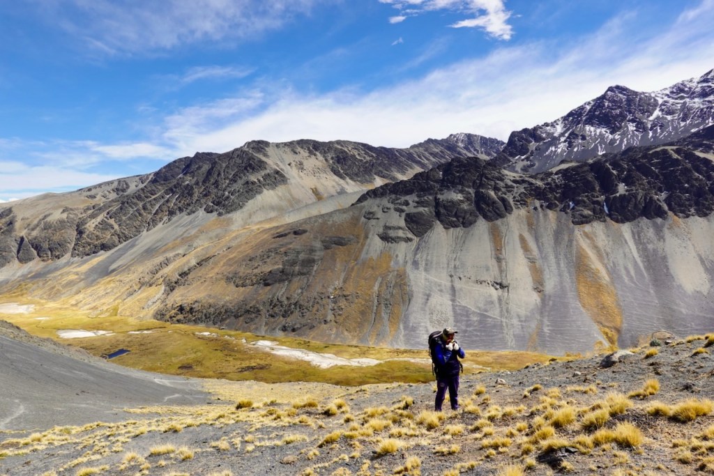 A hiker with a backpack stands on rocky terrain, overlooking a vast valley surrounded by mountains under a blue sky with scattered clouds.