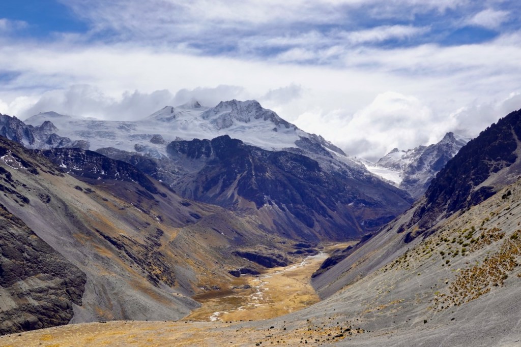 A scenic view of a mountainous landscape featuring rugged peaks, snow-capped summits, and a valley below with dry grass and rocky terrain under a partly cloudy sky.