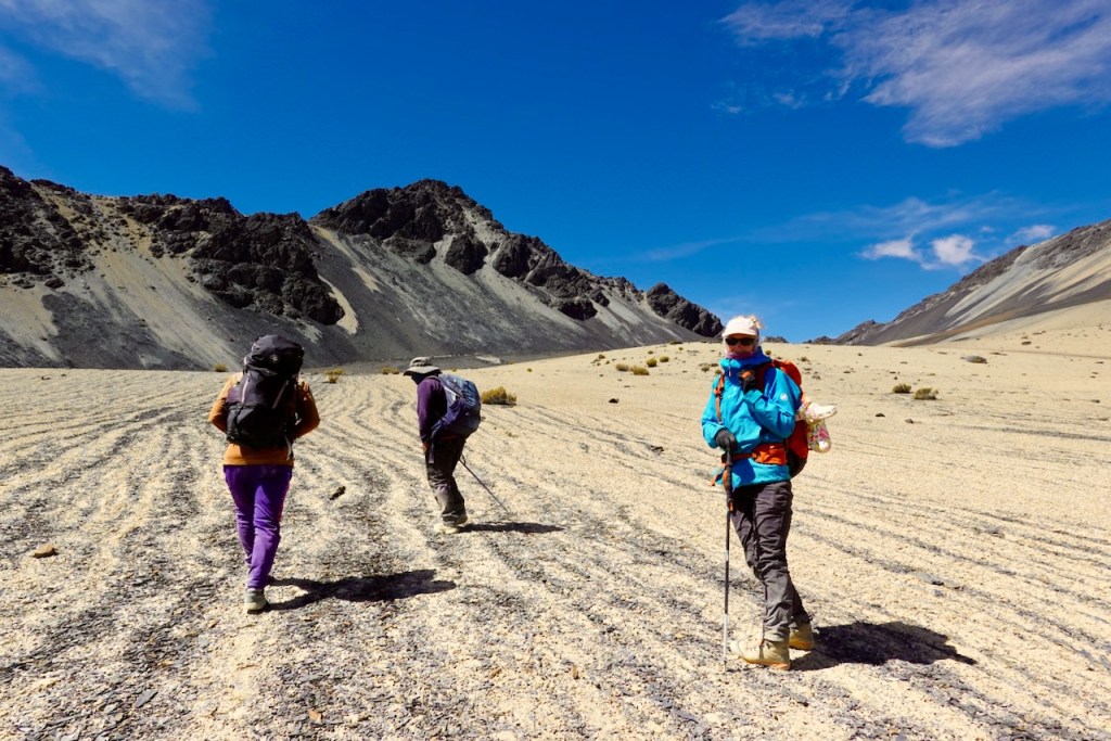 Three hikers trekking through a rocky, sandy landscape with mountains in the background under a clear blue sky.