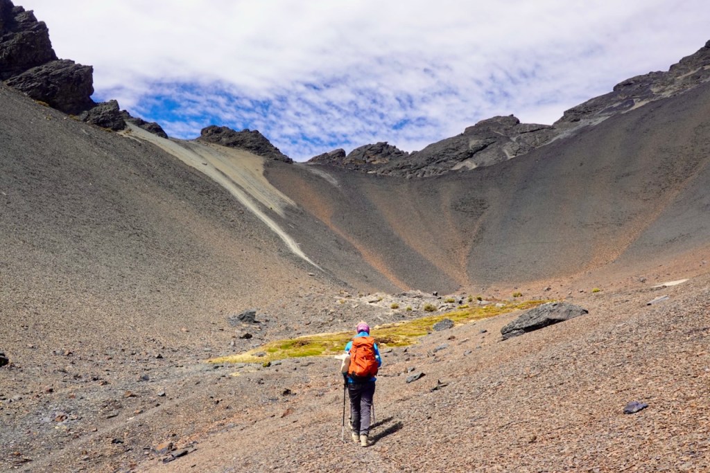 Person hiking on rocky terrain with a steep mountain slope and clouds in the background.
