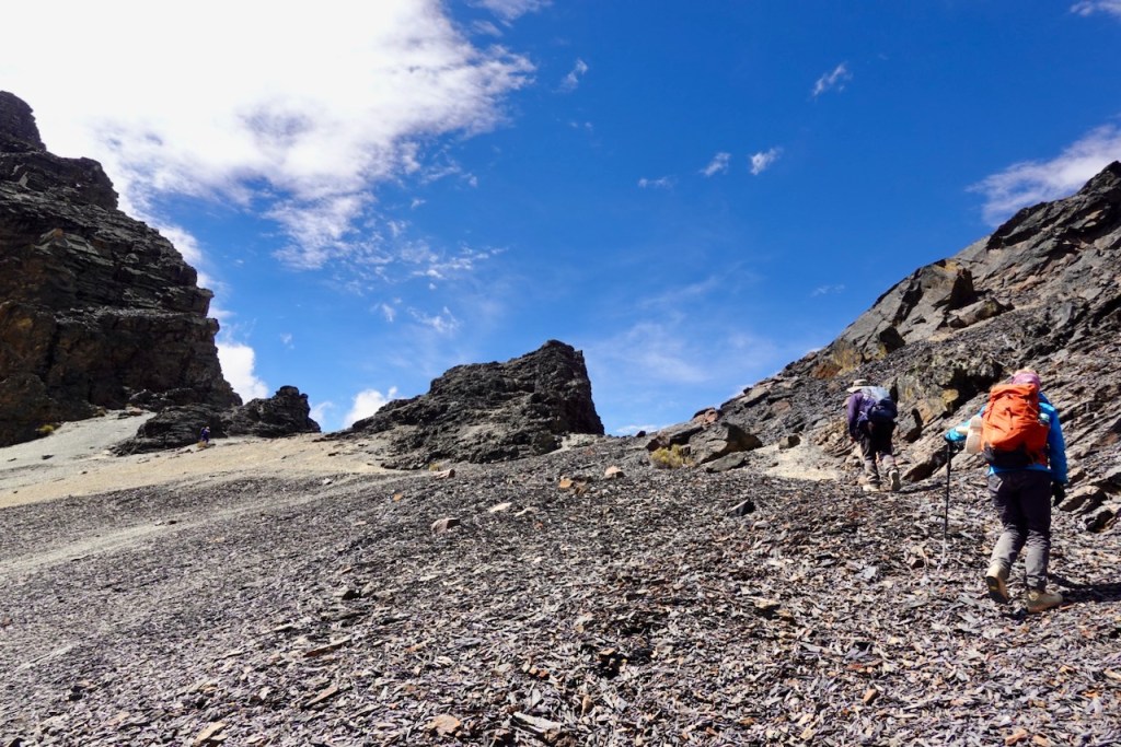 Three hikers ascend a rocky slope under a bright blue sky with scattered clouds.