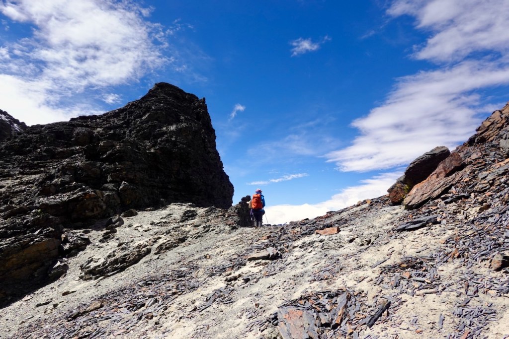 Two hikers walking along a rocky trail with a mountain backdrop and a blue sky.