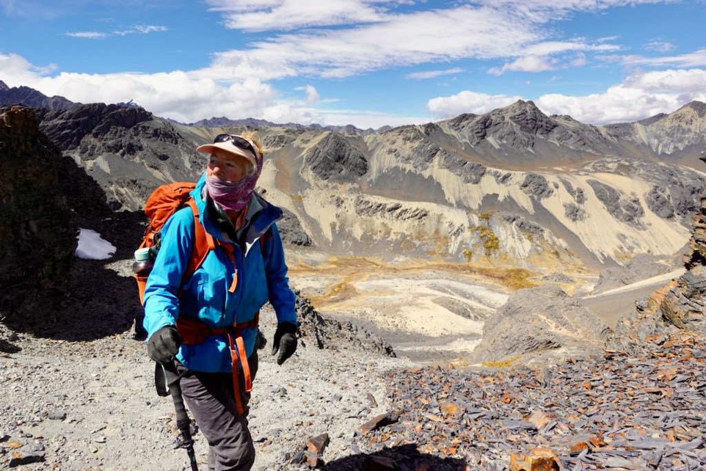 A hiker wearing a blue jacket and an orange backpack stands on a rocky terrain with mountains in the background, looking towards the horizon.