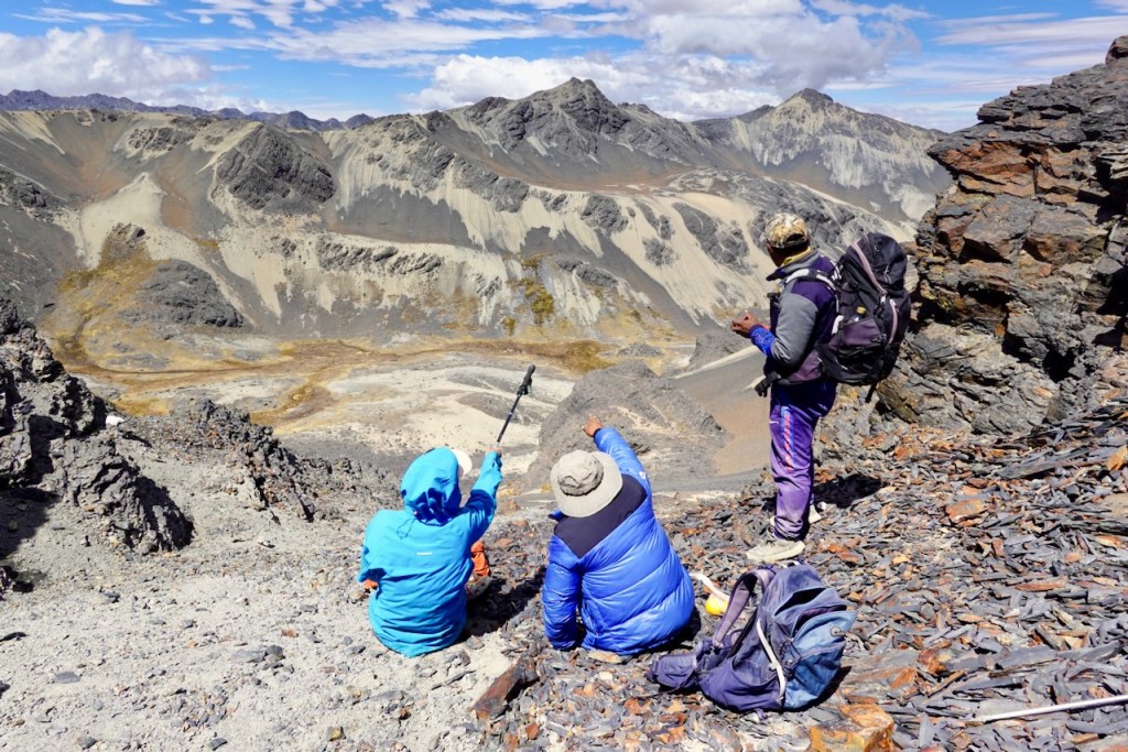 Three hikers sitting on a rocky mountain slope, overlooking a vast valley with rugged terrain and distant peaks under a blue sky.
