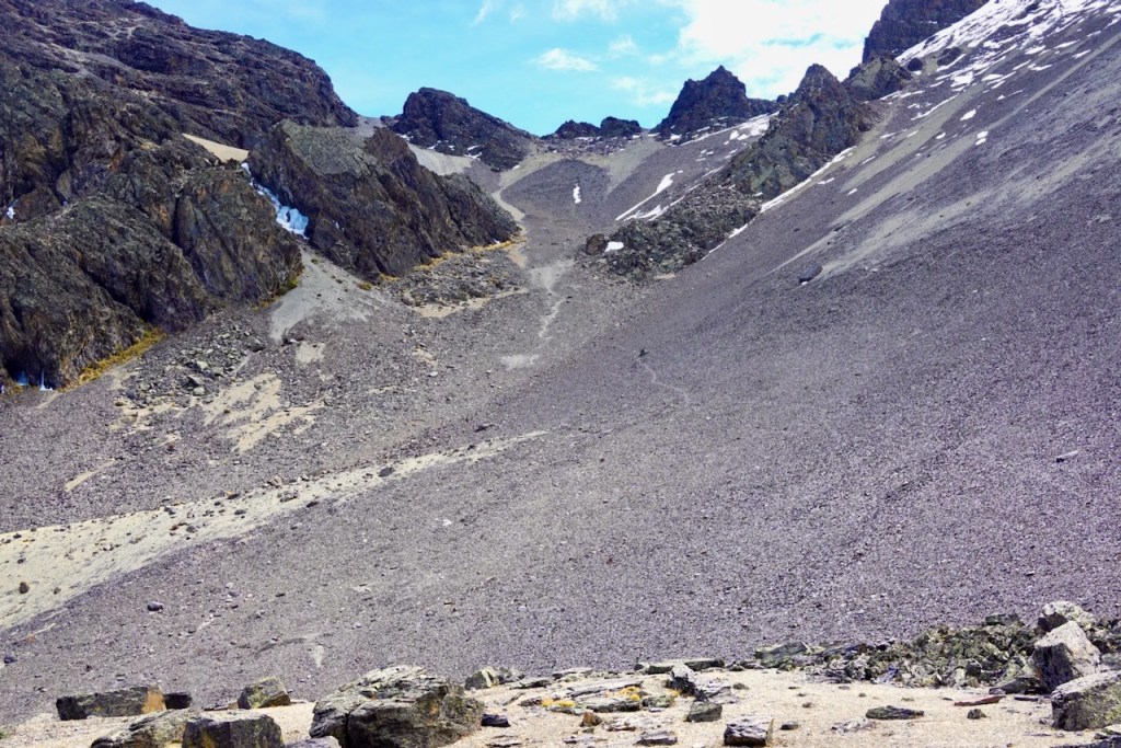 A rocky mountain slope with scattered boulders and a mix of gravel and snow patches under a blue sky.