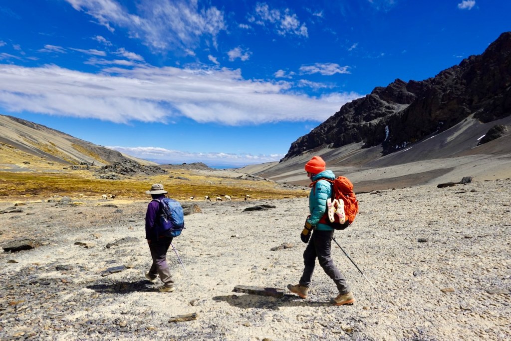 Two hikers walking on a rocky terrain with mountains in the background under a blue sky with clouds.