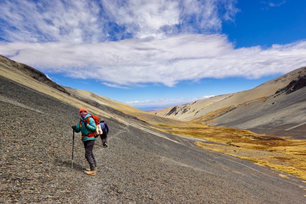 A hiker wearing a colorful jacket and hat climbs a rocky slope in a mountainous landscape, with a clear blue sky and scattered clouds above.