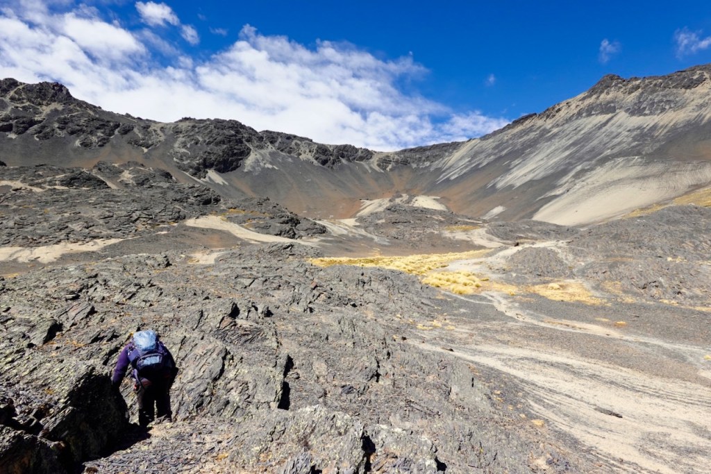A hiker climbing rocky terrain in a mountainous landscape under a blue sky with clouds.