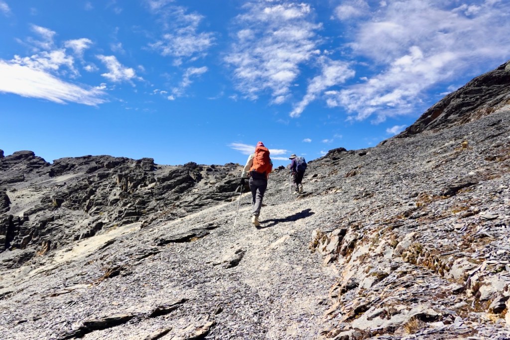 Two hikers ascending a rocky trail under a blue sky with scattered clouds.