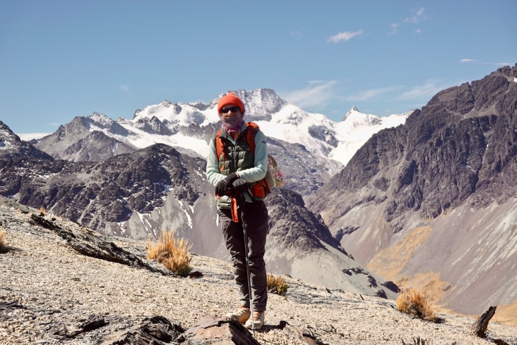 A hiker standing on rocky terrain, wearing an orange beanie and a green vest, with snow-capped mountains in the background under a clear blue sky.