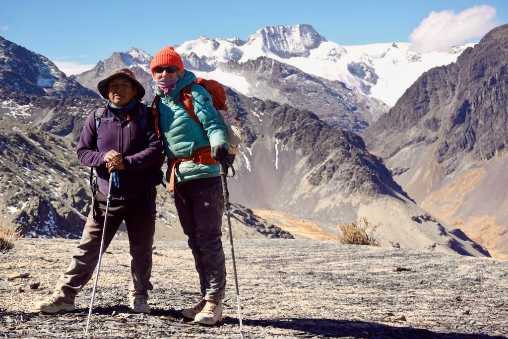 Two hikers posing together on a rocky mountain landscape with snow-capped peaks in the background.