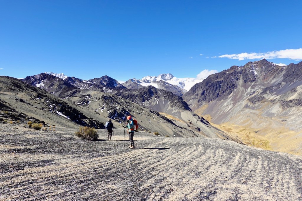 Two hikers walking on a rocky mountain trail with snow-capped peaks in the background and a clear blue sky.