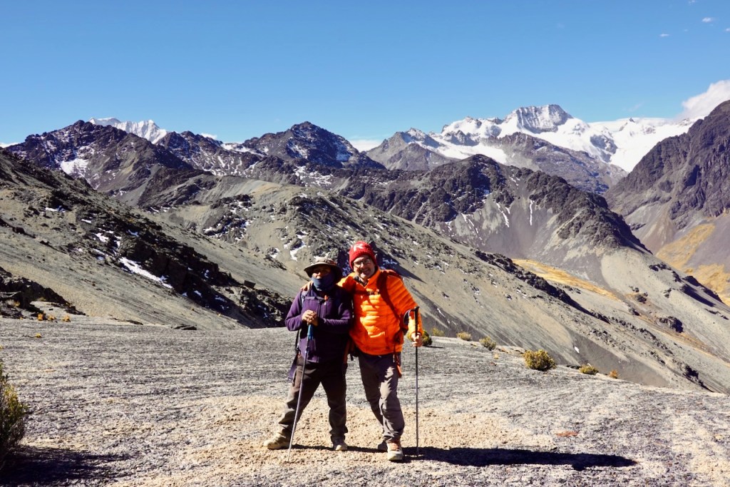 Two hikers standing on a rocky mountain landscape, with snow-capped peaks in the background and a clear blue sky.