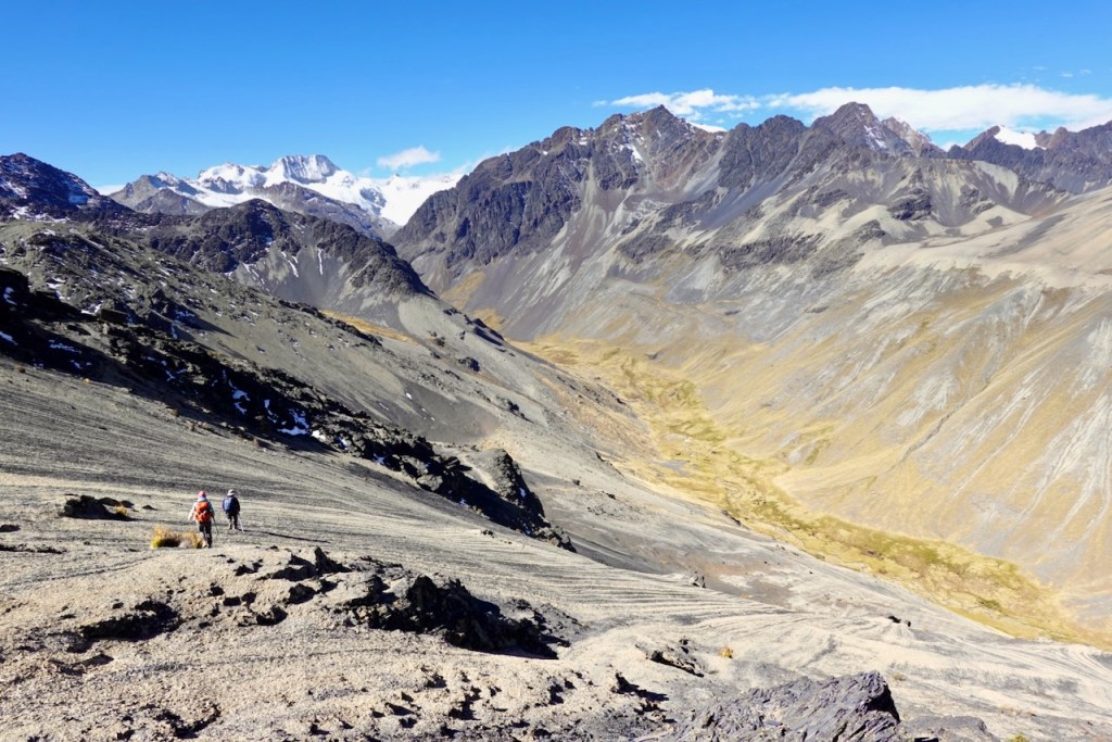 Two hikers walking on a mountainous terrain with snow-capped peaks in the background and a valley below under a clear blue sky.