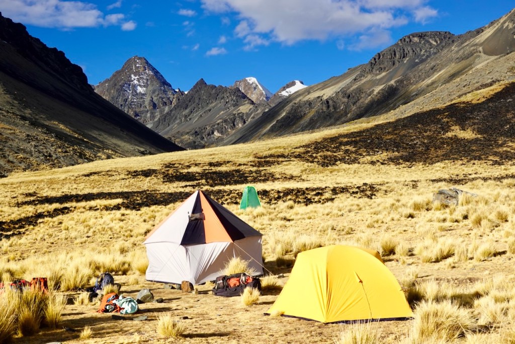 Campsite in a mountainous area featuring tents, grassland, and rocky terrain under a blue sky with clouds.