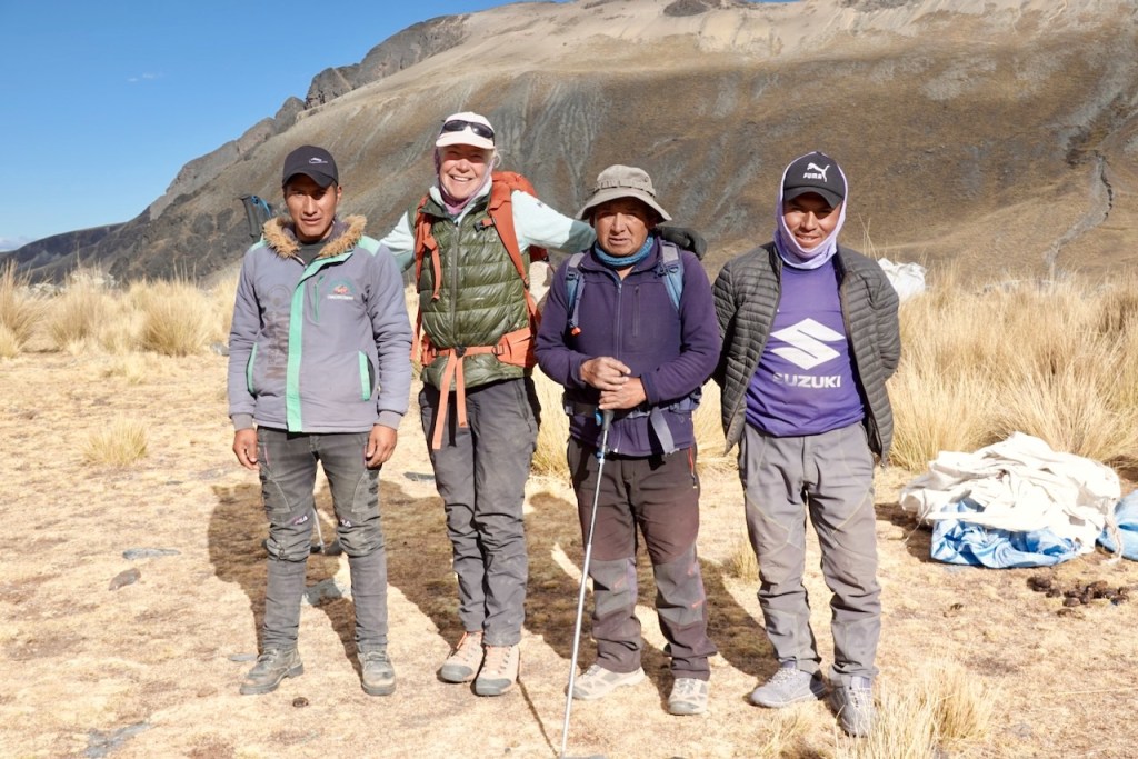 Group of four men standing in a mountainous landscape, dressed in outdoor clothing. They are smiling, with a backdrop of grassland and rocky hills.