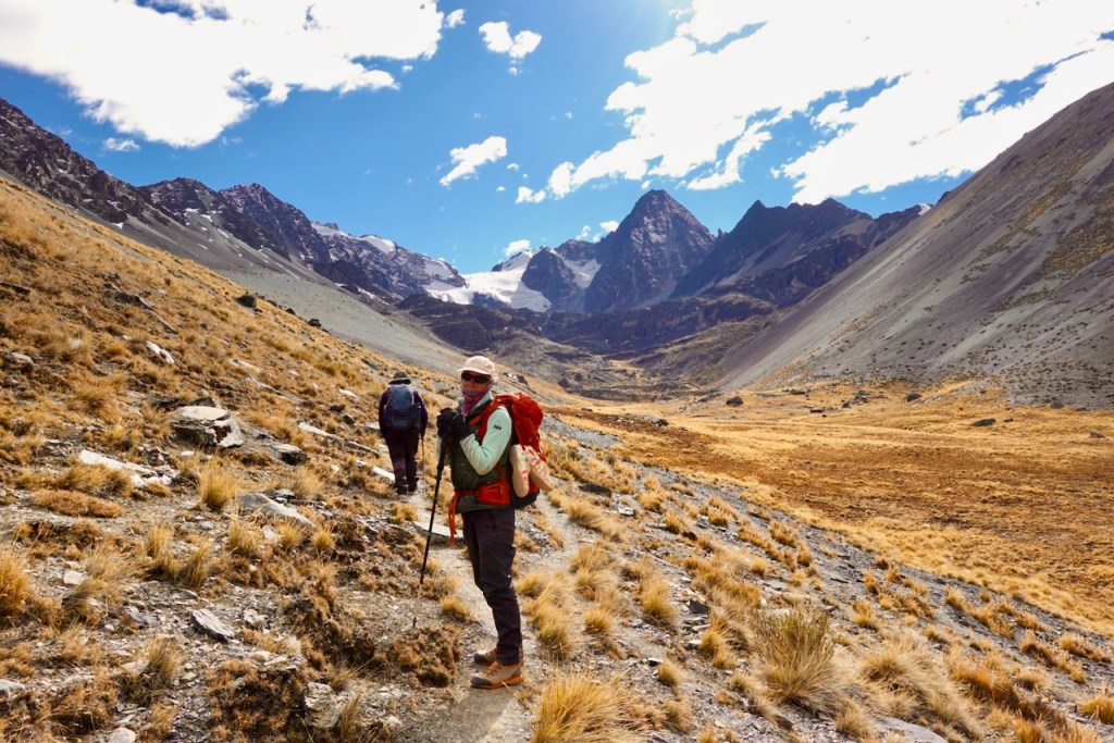 Two hikers trekking through a mountainous landscape with rocky terrain and tall peaks in the background, under a bright blue sky.