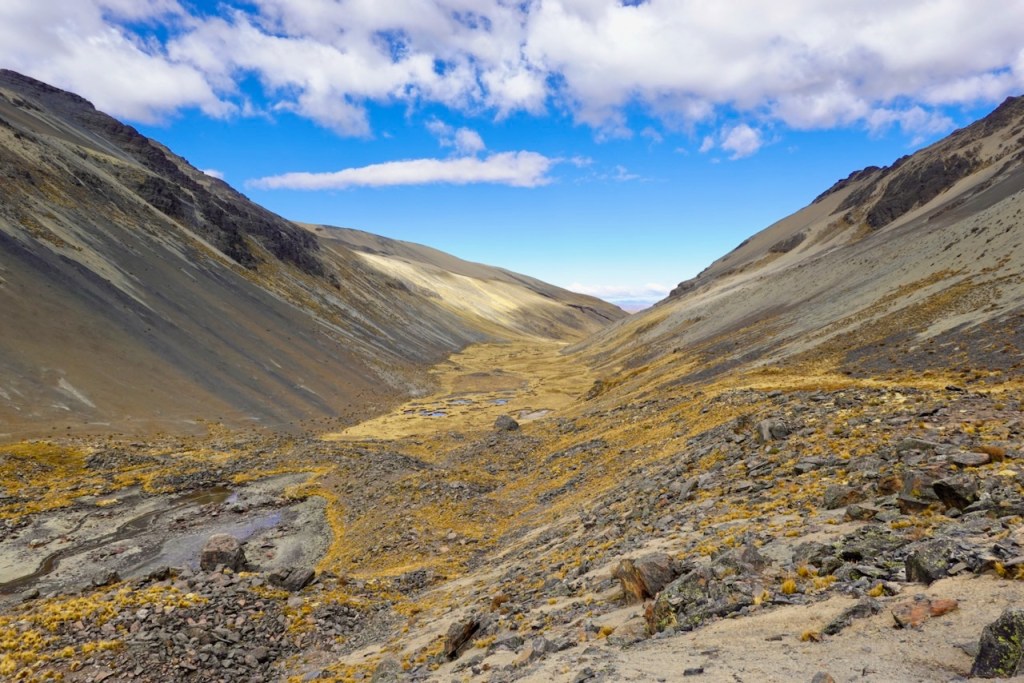 A scenic view of a wide valley surrounded by steep mountains, featuring dry terrain, scattered rocks, and patches of grass under a clear blue sky with some clouds.