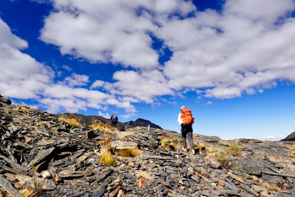Two hikers navigate a rocky trail under a blue sky with fluffy white clouds.