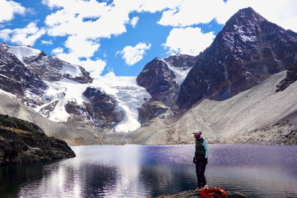 A person standing by a serene alpine lake with snow-capped mountains in the background under a blue sky with scattered clouds.