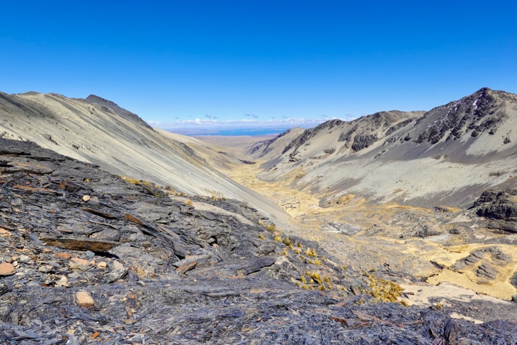 A panoramic view of a rugged mountain landscape featuring steep slopes and a valley below, with clear blue skies in the background.