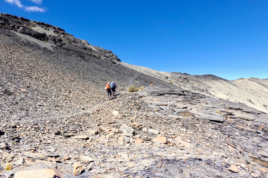 Two hikers walking on a rocky trail with a steep incline under a clear blue sky.