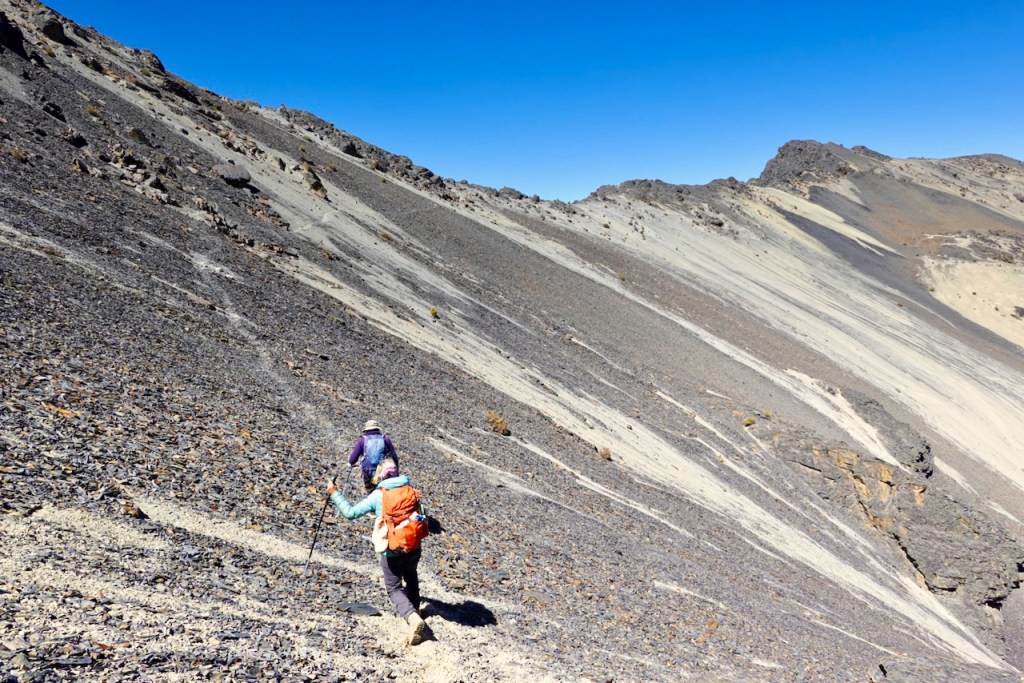 Two hikers ascending a rocky, sloped terrain against a clear blue sky.