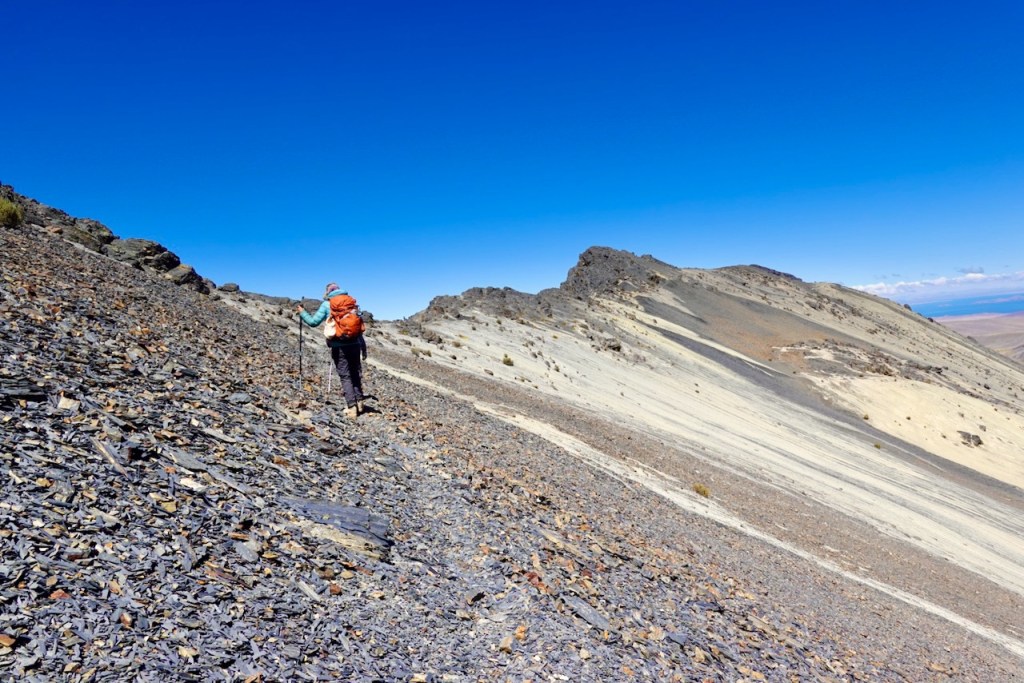 A hiker climbing a rocky slope with an orange backpack under a clear blue sky.