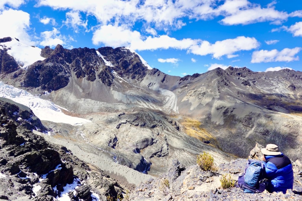 A hiker sitting on a rocky ledge overlooking a vast mountainous landscape with peaks, glaciers, and patches of greenery under a blue sky with clouds.
