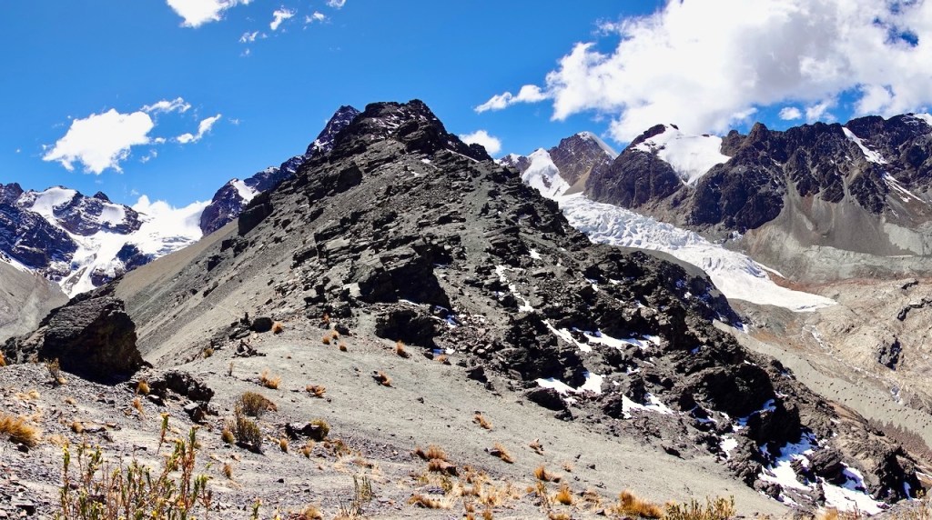A rocky mountain landscape with a steep slope, showing rugged terrain, patches of snow, and a clear blue sky with scattered clouds.