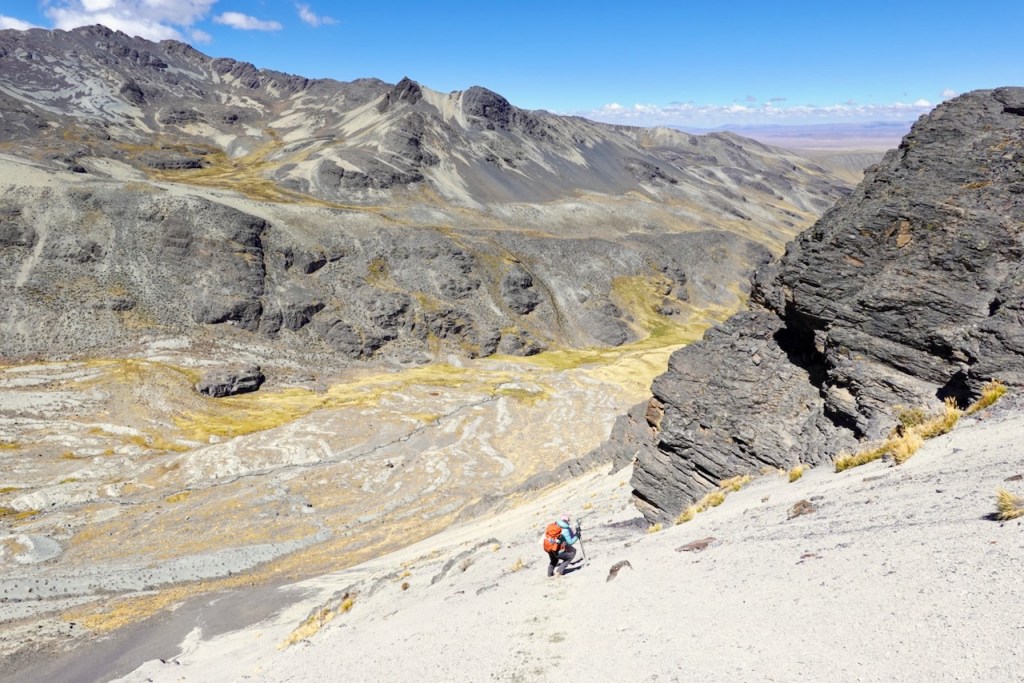 A hiker with a backpack ascends a rocky slope in a mountainous landscape, showcasing rugged terrain and a clear blue sky.