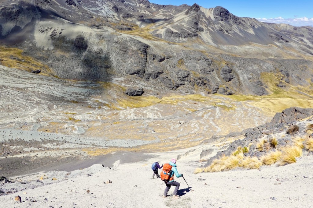 Two hikers trekking up a steep, rocky terrain with a mountainous landscape in the background.
