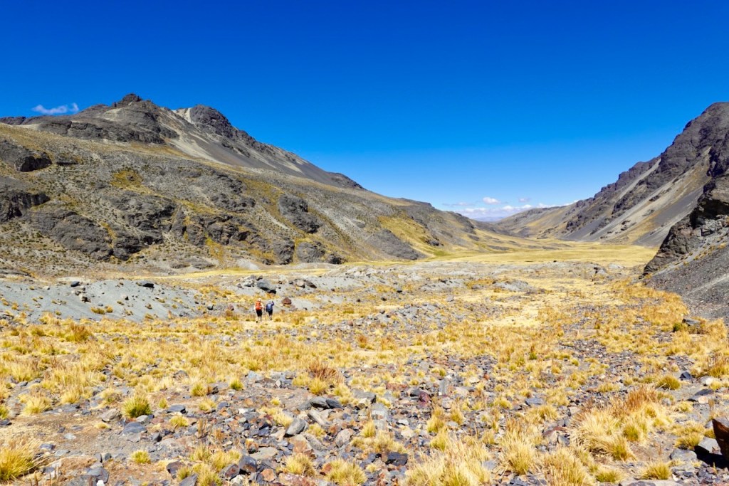 A scenic view of a rugged valley surrounded by mountains, with hikers in the distance walking through rocky terrain and grassland under a clear blue sky.