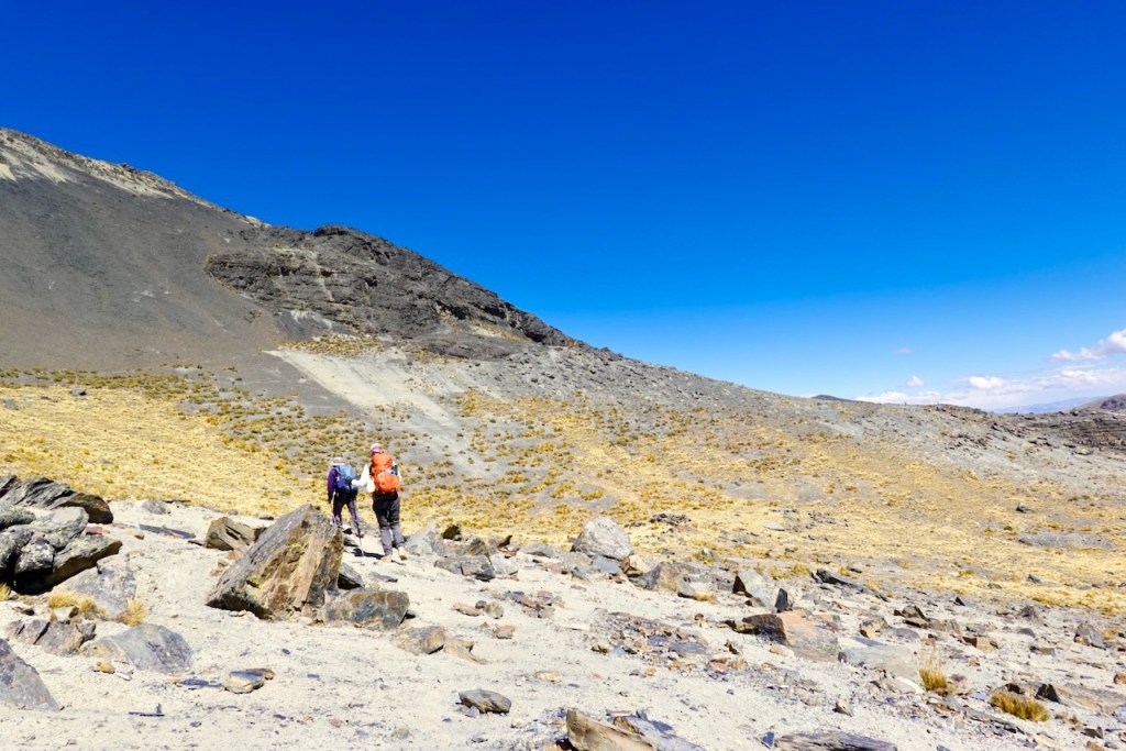 Two hikers with backpacks walking on a rocky trail in a barren, mountainous landscape under a clear blue sky.