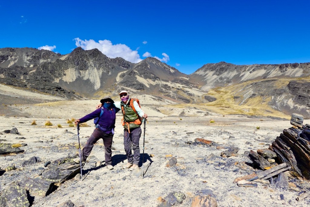 Two hikers standing in a barren mountain landscape, with rugged peaks in the background and clear blue sky.