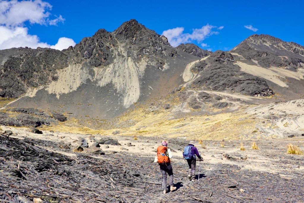 Two hikers walking on a rocky terrain with mountains in the background under a clear blue sky.