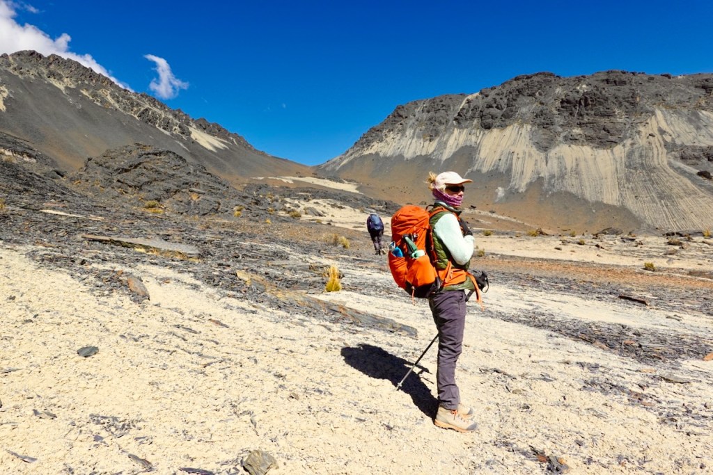 A female hiker in a mountainous landscape, wearing a bright orange backpack and a green jacket, standing on rocky terrain with a sweeping view of the hills and a blue sky.
