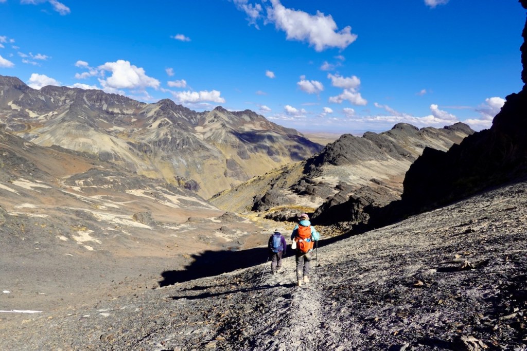 Two hikers walking down a rocky mountain trail with expansive views of rugged peaks and blue sky in the background.