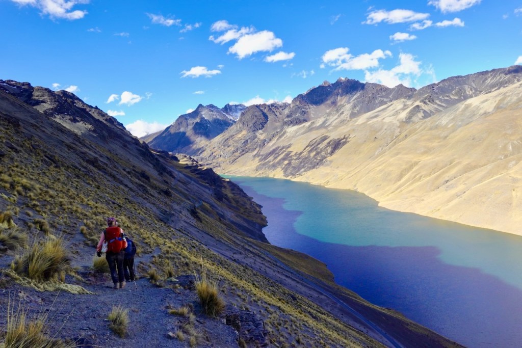 Two hikers walking along a trail beside a turquoise lake, surrounded by mountainous terrain and blue sky with clouds.
