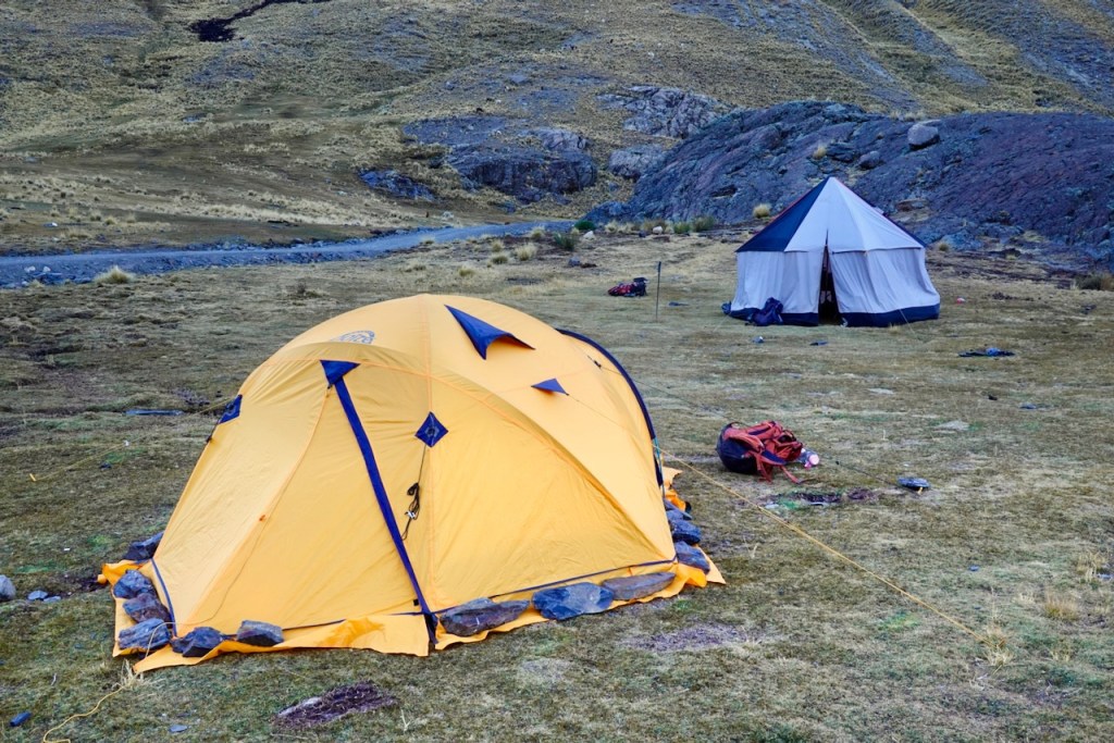A yellow dome tent set up in a grassy campsite with a white and black tent in the background, surrounded by rocky terrain.