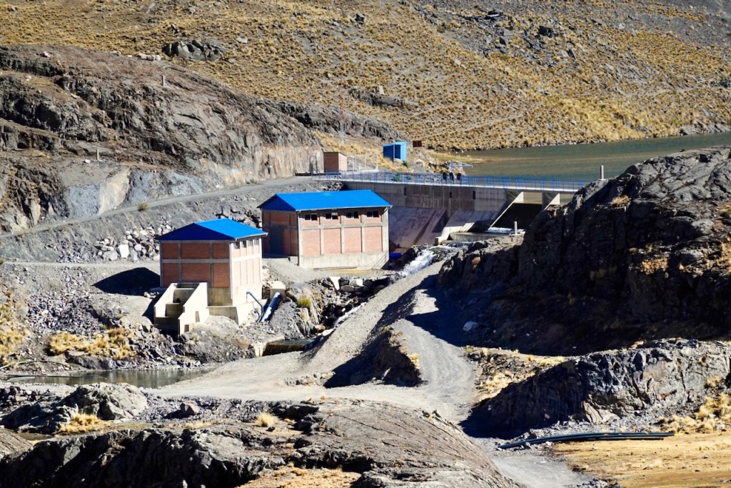 Two buildings with blue roofs situated on a rocky hillside, near a body of water, surrounded by dry grass and rocky terrain.