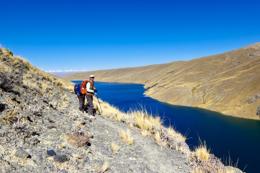 A hiker stands on a rocky path overlooking a blue river surrounded by dry hills under a clear blue sky.