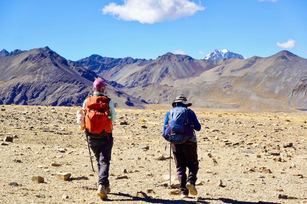 Two hikers walking on a rocky terrain towards mountains, with a clear blue sky in the background.